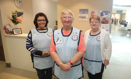 Three women wearing blue aprons standing in front of hospital concierge desk